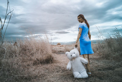 Full length of woman standing with teddy bear on field against cloudy sky