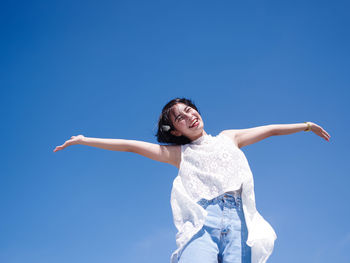 Low angle portrait of happy young woman with arms outstretched standing against blue sky