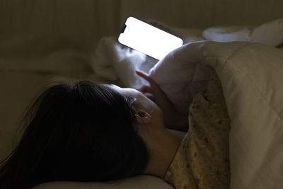 Woman looking at her cellphone under the white blanket, girl lying and rolling in the bed at night