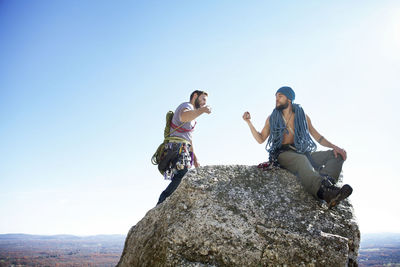 Friends doing fist bump while sitting on rock against clear sky