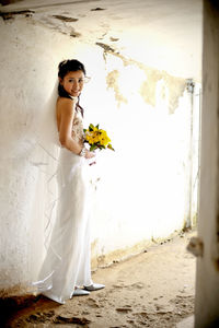 Portrait of smiling bride holding bouquet while standing against wall outdoors