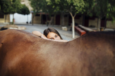 Portrait of young woman leaning on brown horse from behind