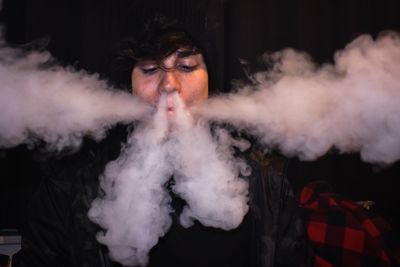 Close-up of young man emitting smoke from nose while standing outdoors at night