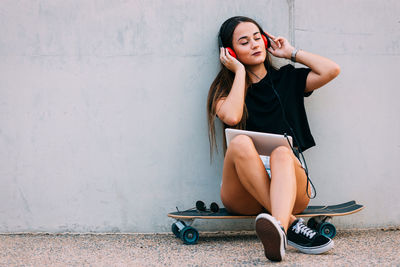 Woman listening music while sitting on skateboard against wall