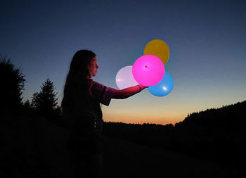 Side view of silhouette woman holding colorful balloons against sky during sunset