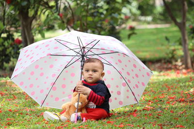 Full length of cute baby boy sitting with umbrella and teddy bear on grass at park