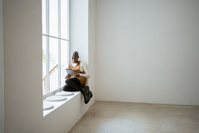 Smiling woman in bib overalls looking at digital tablet while sitting on window sill in living room