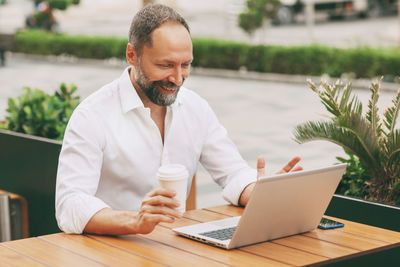 Adult male businessman sitting on the street in a cafe, drinking coffee and working on a laptop.
