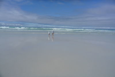 Distant view of siblings walking at beach against sky