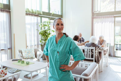 Smiling young elderly care nurse standing with hand on waist at nursing home