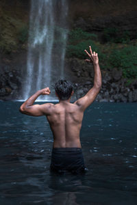 Rear view of shirtless man gesturing while standing in lake by waterfall