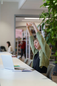 Tired woman adult learner stretching arms with closed eyes while studying in cozy library