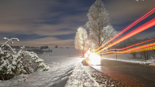 Light trails on road by trees against sky during sunset