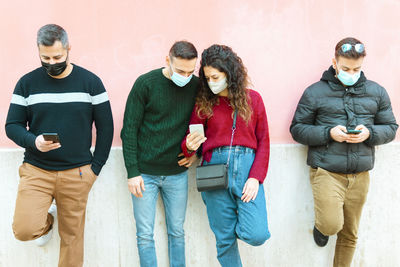 Friends wearing mask using smart phones while standing by wall outdoors