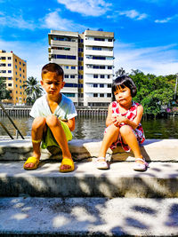 Siblings sitting on promenade in city against blue sky