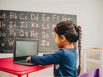 Side view of girl using laptop on table against blackboard