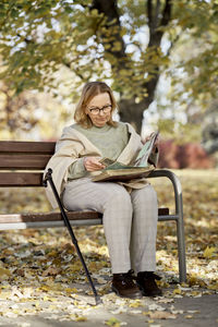 Senior woman looking at photo album sitting with walking stick on bench at park