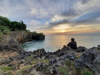 Man sitting on rock by sea against sky during sunset