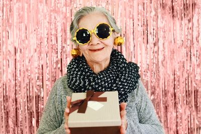 Portrait of smiling woman wearing sunglasses holding gift while standing against wall