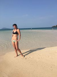 Portrait of beautiful woman in bikini with hands on hip standing at beach against blue sky