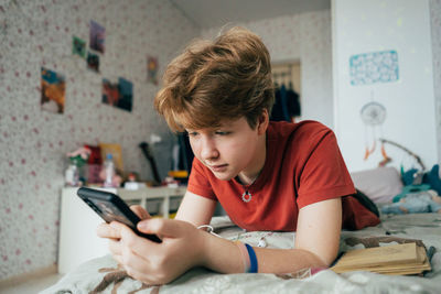 A teenage girl lying on a bed in a room uses a mobile phone for online communication.