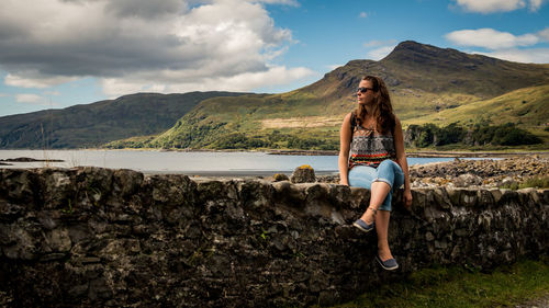 Young woman sitting on retaining wall at lakeshore against mountains