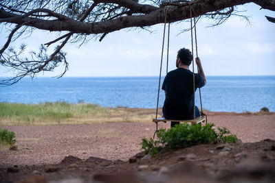 Rear view of man sitting on rope swing against sea