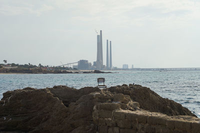 Chair on rocks, factory,  sea, against sky