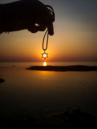 Silhouette hand holding necklace over sea against sky during sunset