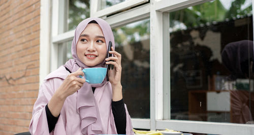 Woman wearing hijab holding coffee cup while talking over mobile phone at cafe