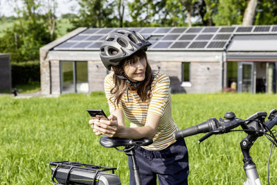 Smiling woman with bicycle and cell phone on a meadow in front of a house