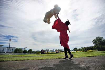 University student throwing teddy bear against sky during graduation