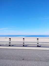 Empty road by fence against sea and blue sky