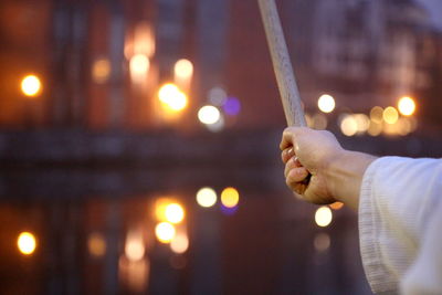 Cropped image of man practicing martial arts with sword against illuminated canal in city during sunset