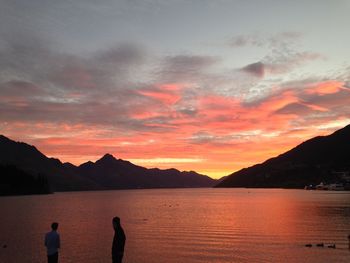 Men standing by lake against dramatic sky during sunset