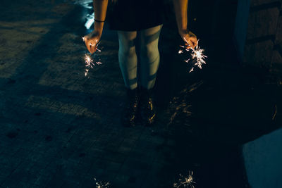 Low section of woman holding sparklers while standing on footpath at night