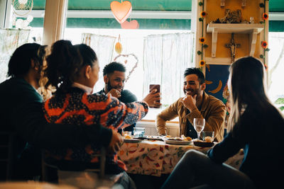 Young woman showing smart phone to smiling man sitting at restaurant during brunch party