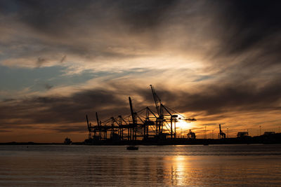 Silhouette cranes at harbor against sky during sunset