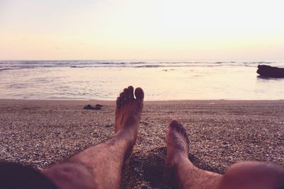 Low section of man relaxing at beach against sky during sunset