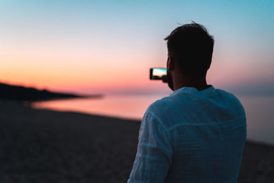 Rear view of man photographing sea against sky during sunset