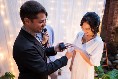 High angle of multiethnic newlywed couple smiling while standing near arch decorated with curtain and light bulb garlands while exchanging rings during wedding celebration