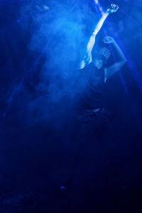 Full body of unrecognizable muscular male enthusiastically dancing in dark room in vapour and beams of bright blue lamp
