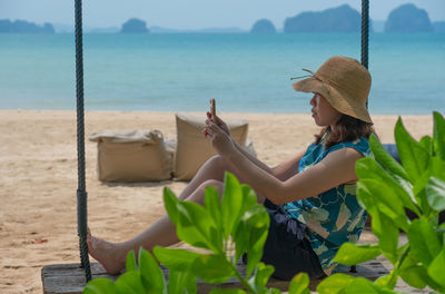 Young woman sitting on swing near beach and using smartphone to take photo during summer vacation