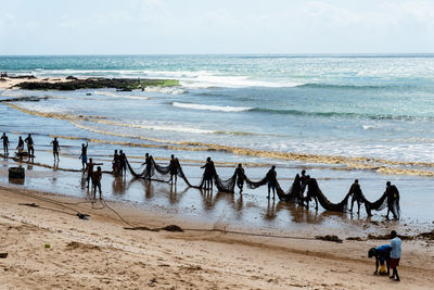 Several fishermen are seen walking in a line and carrying a large fishing net. 