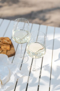 Two glasses of white wine and a wooden plate with cheese and nuts on a white table outdoors.