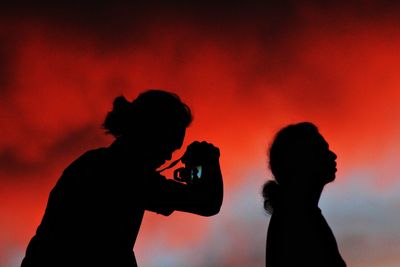 Silhouette man photographing woman against orange sky