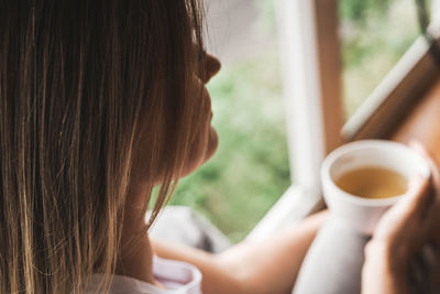 Woman drinking a cup of hot tea or coffee while sitting on the windowsill in the morning