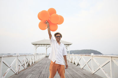 Portrait of man holding balloons while standing on pier against sky