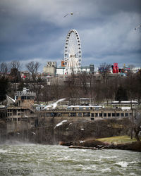 Ferris wheel by river against buildings in city