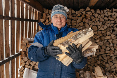 Senior farmer in a warm hat and jacket holds firewood against the background 